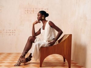 Imagem com bege claro, com uma uma mulher de cabelo preso em coque sentada em uma cadeira de madeira marrom, ela esta usando um vestido longo branco e uma sandalia de salto baixo com amarraçõa.