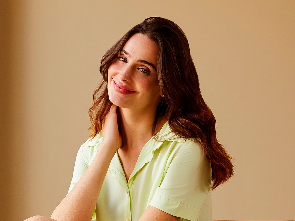 Imagem com fundo marrom claro, com uma mulher de cabelo longo castanho e vestindo uma blusa verde claro.