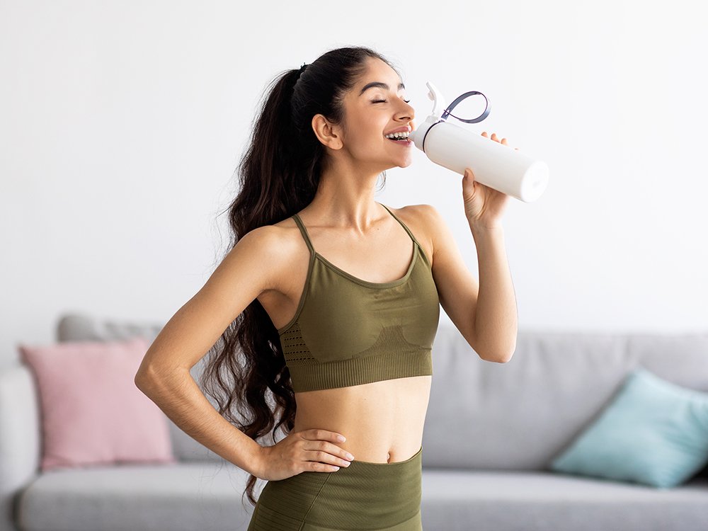 Mulher usando um tópmde academia verde militar bebendo água em uma garrafinha branca